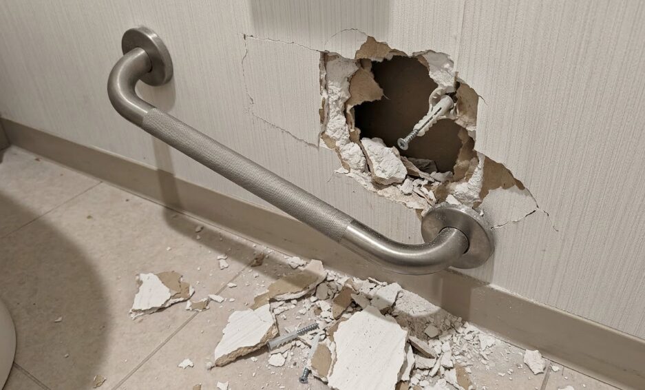 石膏ボードの壁から外れかけた手すりと、崩れた壁の破片。DIYでの不適切な取り付けによる危険性を警告する画像。