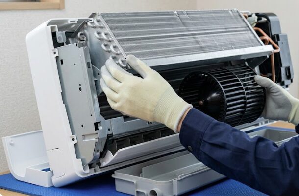 ドレンパンが外されたエアコンで、日本人の男性作業員が熱交換器を左側から慎重に持ち上げ、シロッコファンを斜め下に引き抜く様子。内部の繊細な構造が鮮明に写っている。