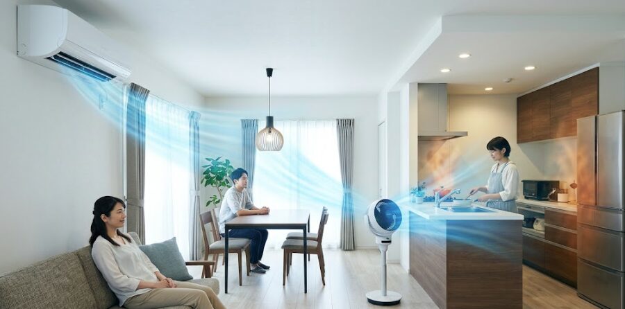 広いLDK全体を映した写真。左のリビング側の壁上部にあるエアコンから吹き出す冷風が、中央のサーキュレーターに到達。サーキュレーターから生まれた強い気流がキッチンまで届き、調理中の女性（日本人）が涼しげにしている。ダイニングライトに風が直撃していない様子も確認できる。