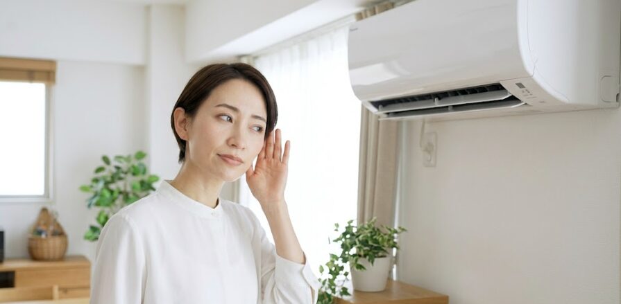 リビングで動いているエアコン室内機の方を向き、耳をそばだてて異音を確認している日本人女性