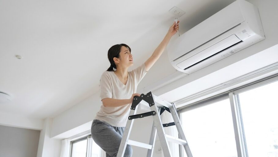 天井に設置された高いコンセントにプラグを抜くため、脚立に登って手を伸ばす日本人の住人。抜き差しのしにくさを描写。