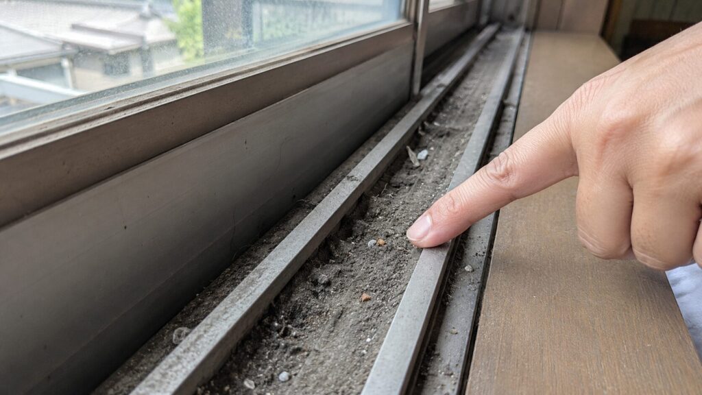 汚れやホコリ、ゴミが大量に溜まったアルミサッシのレールの溝。動きが重くなる原因を指で指し示している。