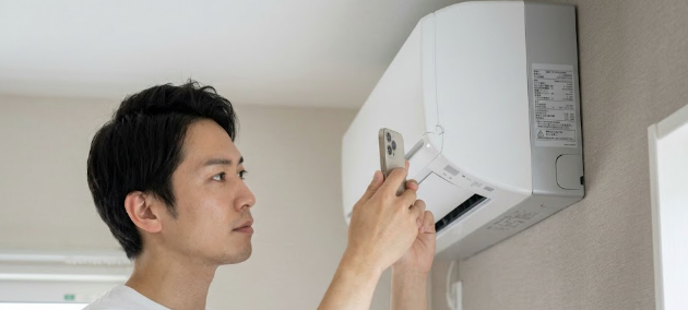 リビングルームで、日本人男性が脚立に乗り、エアコン本体の側面に貼られた型番シールをスマートフォンで撮影している様子。