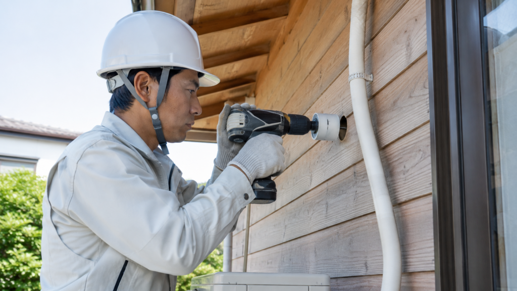 木造住宅の壁に、エアコン配管を通すための穴を標準工事の範囲内で丁寧に開ける日本人技術者。